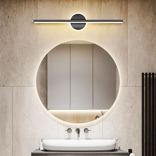 "A modern black wall lamp with a cylindrical bar intersecting a circular base, mounted above a round mirror in a contemporary bathroom with a white vessel sink."