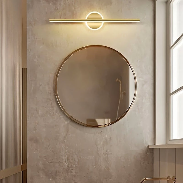 "A modern gold wall lamp with a cylindrical bar intersecting a circular base, mounted above a round mirror in a contemporary bathroom with textured walls."