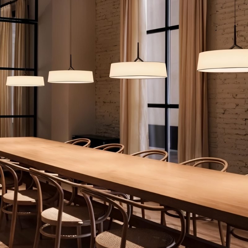 "Multiple pendant lights with wide, white fabric shades illuminate a long table in a commercial space with large windows."