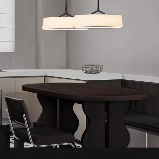 "Two pendant lights with wide, white fabric shades hang above a dark wood table in a modern kitchen."