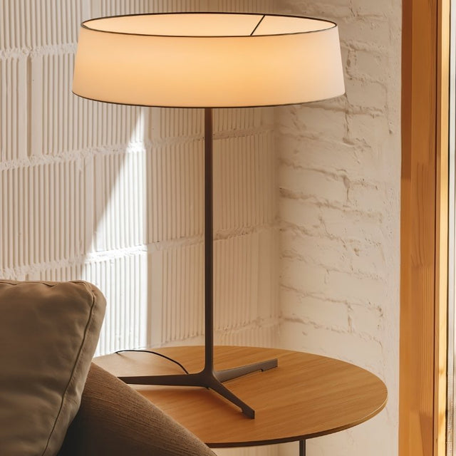 "A contemporary table lamp with a fabric drum shade sits on a round side table next to a sofa in a modern living room."