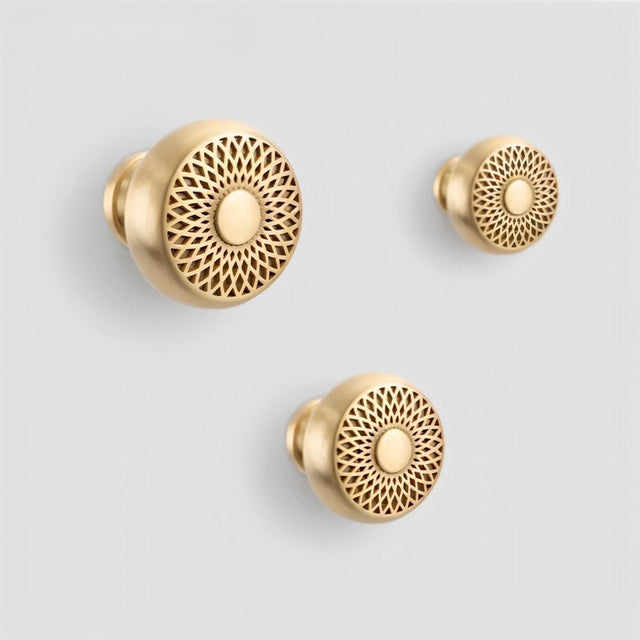"Three gold brass geometric knobs with a lattice and sunburst design on a light gray background."