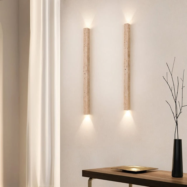"Pair of minimalist rectangular travertine wall lamps mounted on a living room wall, providing soft ambient up and down lighting, with a curtain and a decorative vase in the background."
