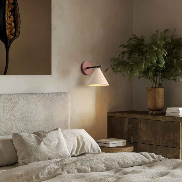 "A modern wall lamp with a conical travertine shade and a black arm, mounted on a wooden backplate in a bedroom, providing a soft, warm glow."