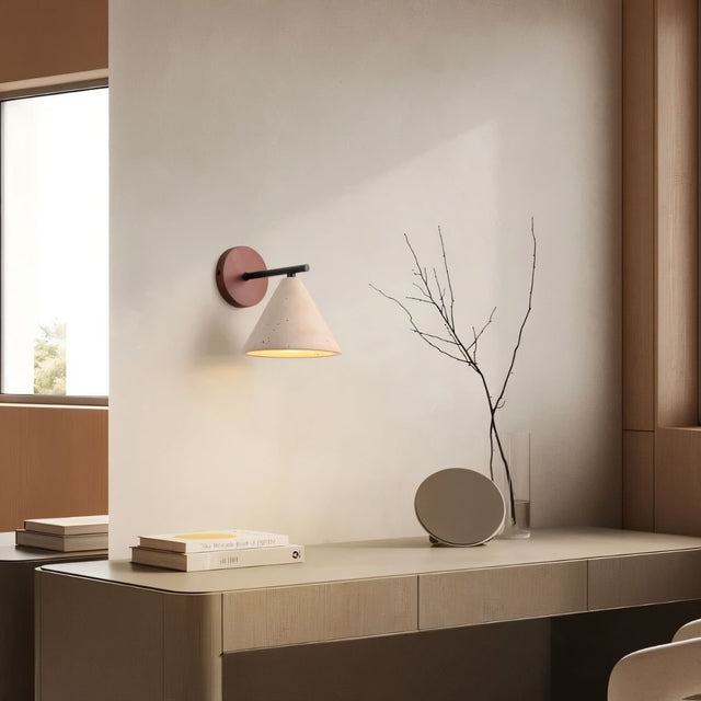 "A modern wall lamp with a conical travertine shade and a black arm, mounted on a wooden backplate above a desk, providing a soft, warm glow to the workspace."