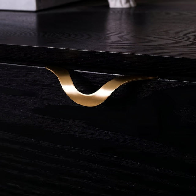 "A brass drawer pull with an elegant, wavy design mounted on a black dresser, adding a luxurious touch to a modern interior."