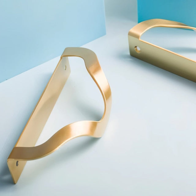 "Brass drawer pulls with an elegant, wavy design displayed on light blue and white geometric shapes, showcasing their modern aesthetic."