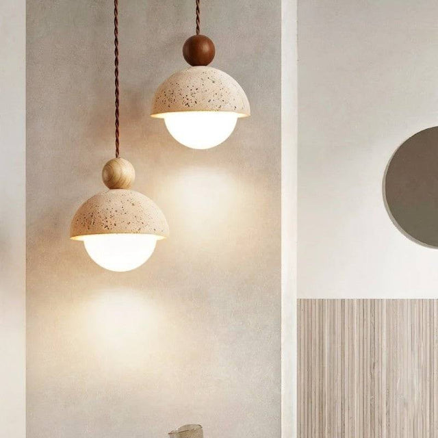 "Two Palermo pendant lights, crafted from yellow travertine with wood accents, hang side-by-side against a textured wall, showcasing their minimalist design and warm light."