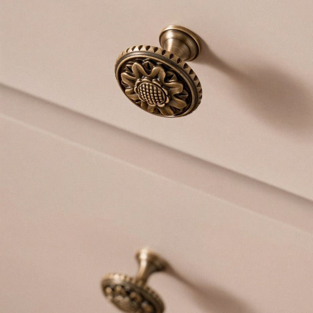 "Antique brass floral knob with detailed floral motif and textured border on a light cabinet drawer."