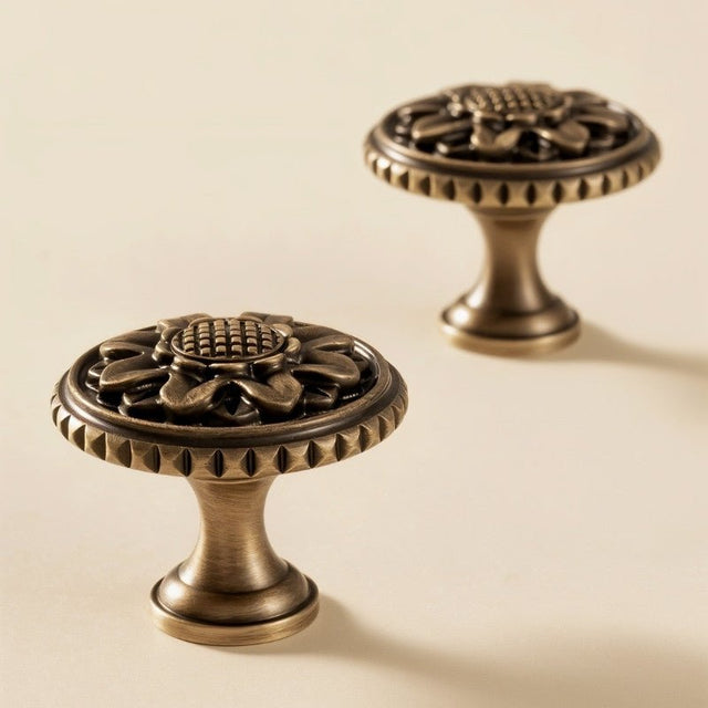 "Close-up of two antique brass floral knobs with detailed floral motif and textured border on a cream surface."
