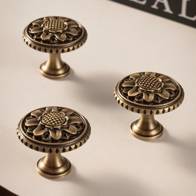 "Three antique brass floral knobs with detailed floral motif and textured border on a light surface."