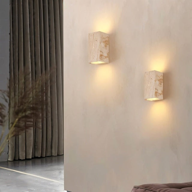 "A pair of modern, cube-shaped wall lamps made of yellow travertine, mounted on a textured wall in a hallway, emitting a soft, ambient glow."