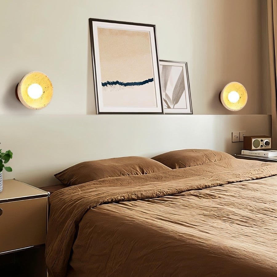 "Pair of travertine wall lamps with circular wooden backplates, mounted on a bedroom wall to provide soft, ambient lighting."