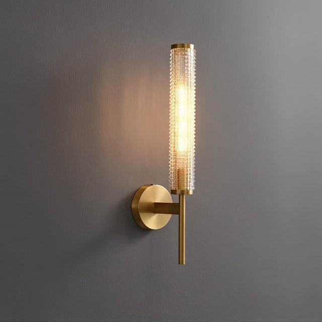 "A modern brass wall lamp with a textured cylindrical glass shade emitting a warm light on a dark gray wall."