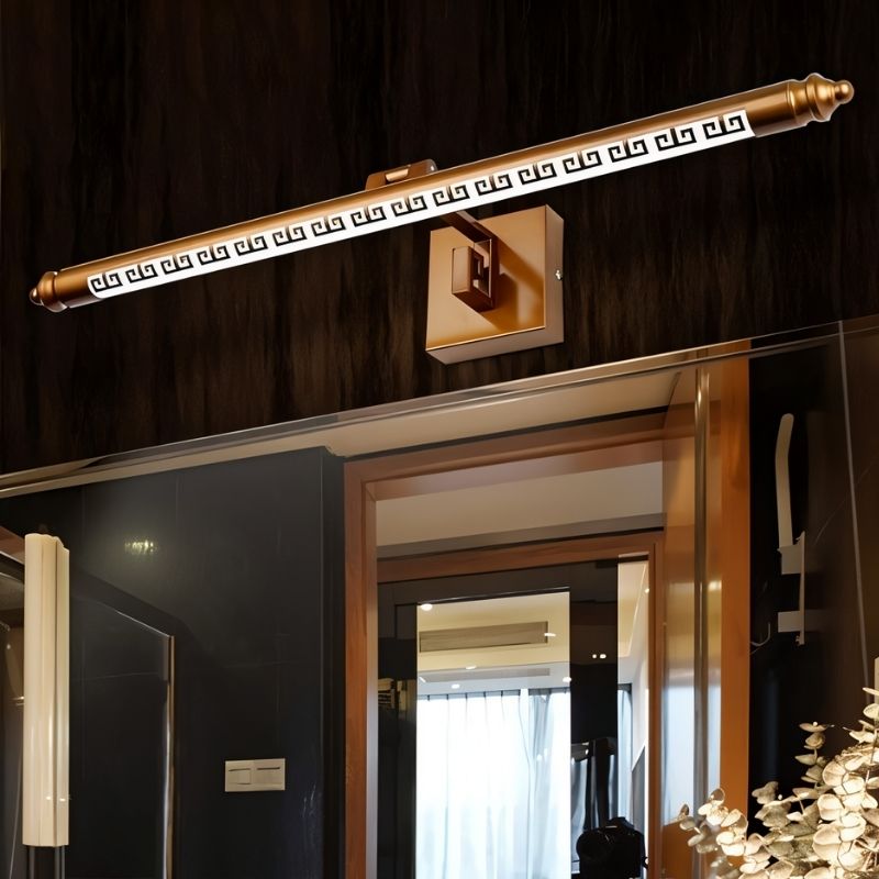"An illuminated gold wall lamp with a Greek key pattern mounted above a bathroom mirror, showing the light reflecting on the mirror and surrounding surfaces."