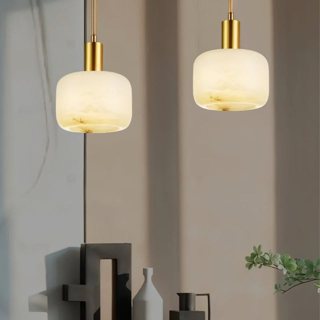 "Two modern pendant lights, each featuring a cylindrical alabaster shade with natural veining and brass accents, hanging against a neutral background."
