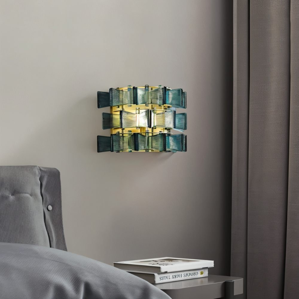 "Iron Salane wall lamp with layered blue glass panels, mounted on a grey wall beside a bed with a book on a nightstand."