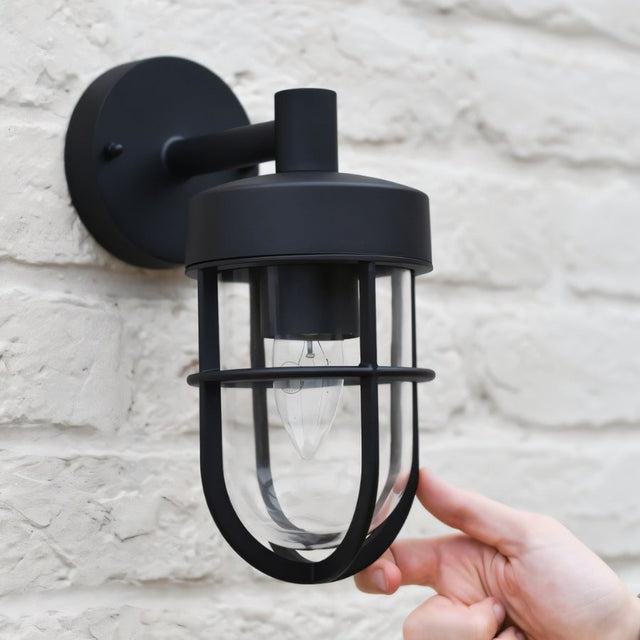 "Hand adjusting a black Siam outdoor wall lamp with a cylindrical glass casing, mounted on a textured brick wall."