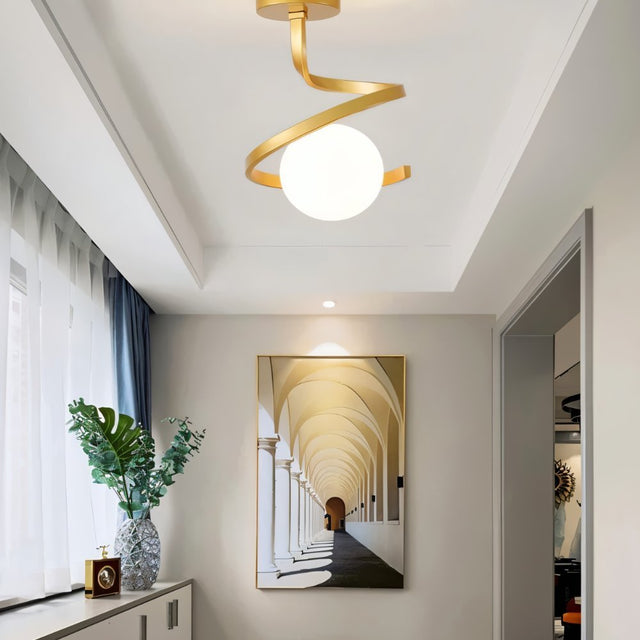 "A modern gold ceiling light with a spiral frame and frosted glass globe in a hallway with artwork and a plant."