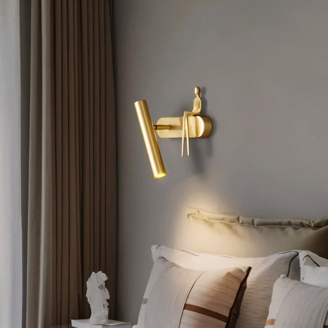 "A gold bedside reading lamp with a figurine on a wall above a bed with pillows."