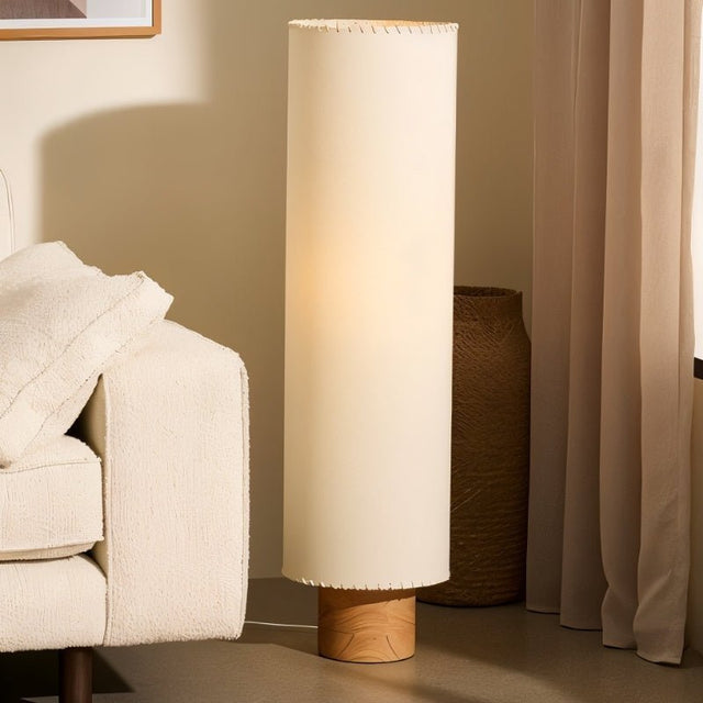 "Svetlina floor lamp placed in a cozy living room corner with a cylindrical fabric lampshade and wood base, offering warm illumination."