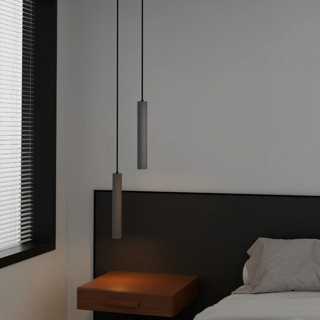 "Two gray cylindrical pendant lights hanging above a nightstand in a bedroom with a window and blinds."