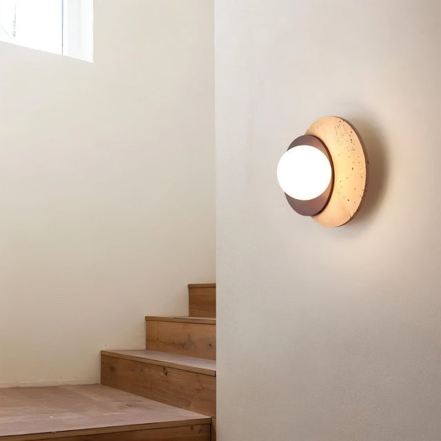 "A wabi-sabi wall lamp featuring travertine and wood, mounted on a wall near a wooden staircase, casting a soft, ambient glow."