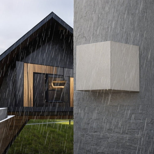 "Tine Wall Lamp mounted on a gray outdoor wall in the rain, featuring a minimalist geometric design made of durable gypsum and aluminum, with upward and downward light beams."
