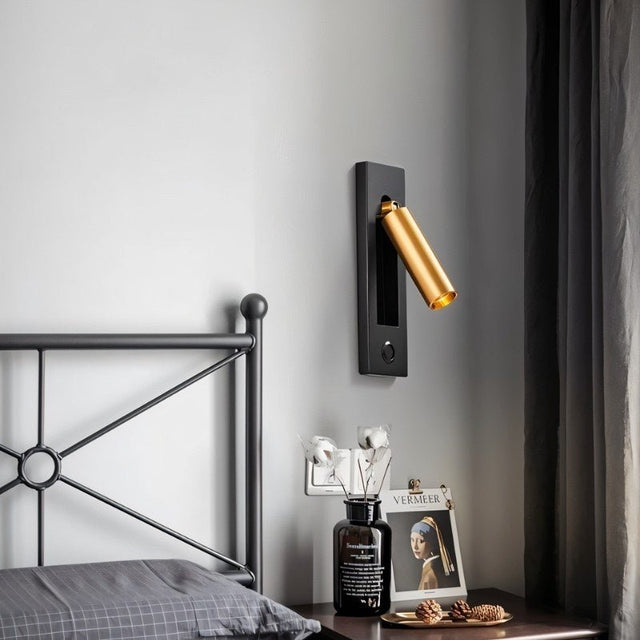 "A black and gold wall lamp with a rectangular base and a cylindrical spotlight, shown mounted on a light gray wall above a nightstand with a vase, pine cones, and artwork featuring Vermeer's 'Girl with a Pearl Earring', next to a metal bed frame with a gray pillow."