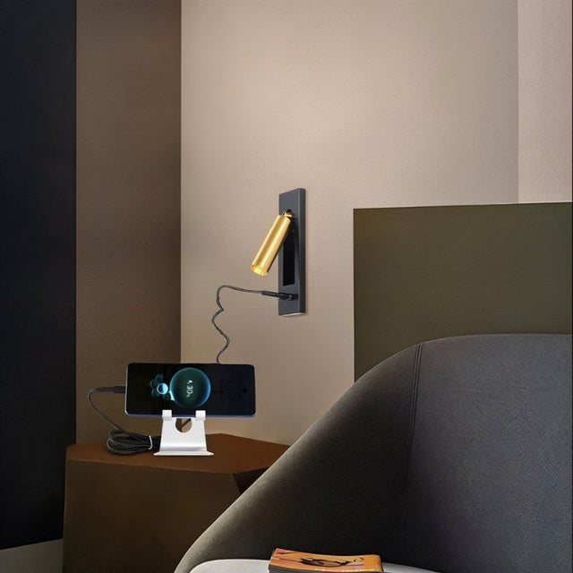"A black and gold wall lamp with a rectangular base and a cylindrical spotlight, shown mounted on a light gray wall above a nightstand with a phone charging on a stand, next to a chair."