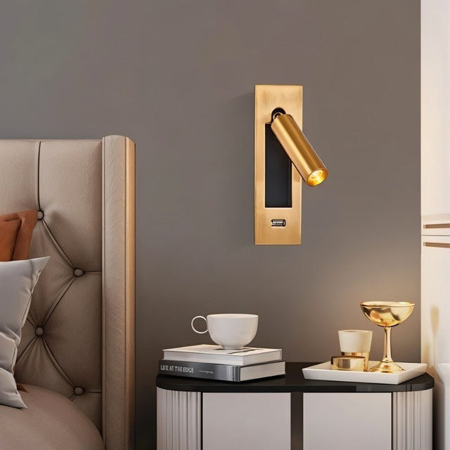 "A gold wall lamp with a rectangular base and a cylindrical spotlight, shown mounted on a gray wall above a nightstand with a book, cup, and gold decorative items, next to a bed with a tufted headboard."