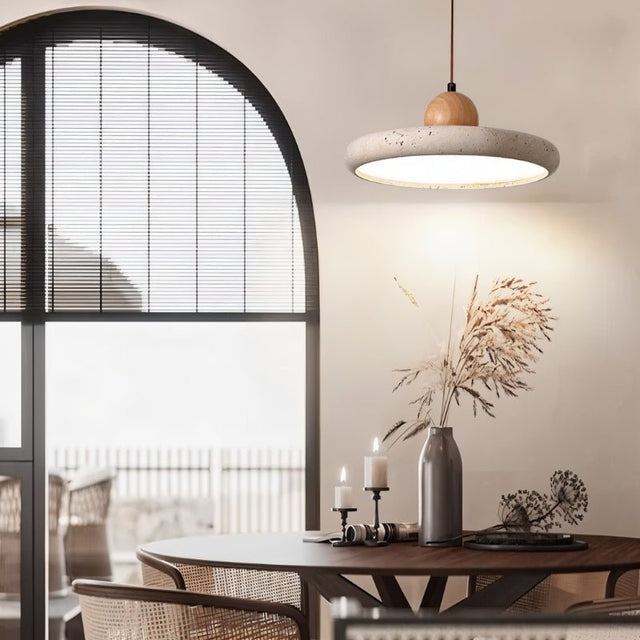 "Modern pendant light with a circular travertine shade and wooden sphere, illuminating a round dining table with decorative items and a large window with blinds."