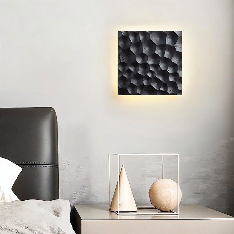 "Black travertine wall lamp with hexagonal pattern, backlit glow, mounted above a bedside table with decorative objects."