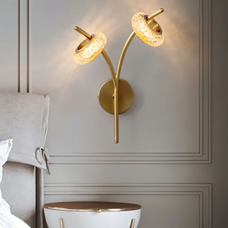 "A modern wall lamp with two textured glass shades and a gold metal frame mounted beside a bed."