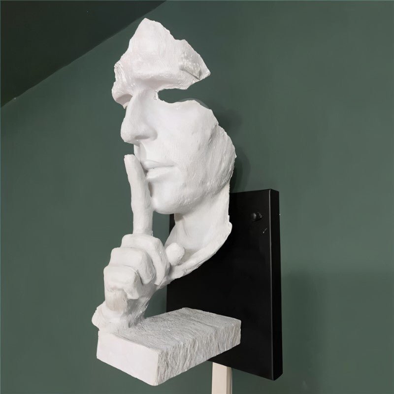 White sculptural face wall lamp with hand gesture mounted on black bracket against green wall.