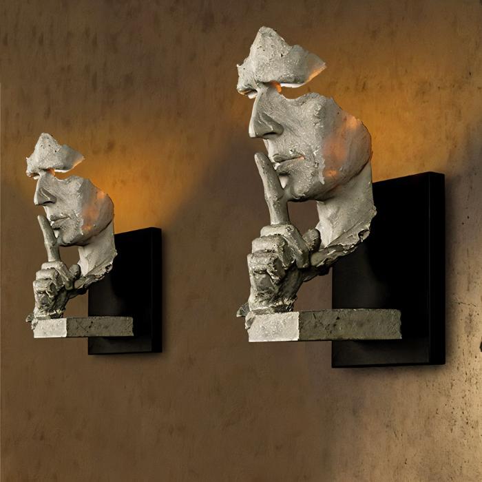 Two concrete sculptural face wall lamps with black mounting brackets against a textured brown backdrop.