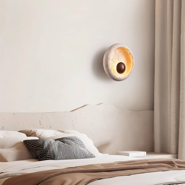 "A minimalist wall lamp with a circular travertine frame and warm wooden centerpiece serving as a sconce in a modern bedroom."