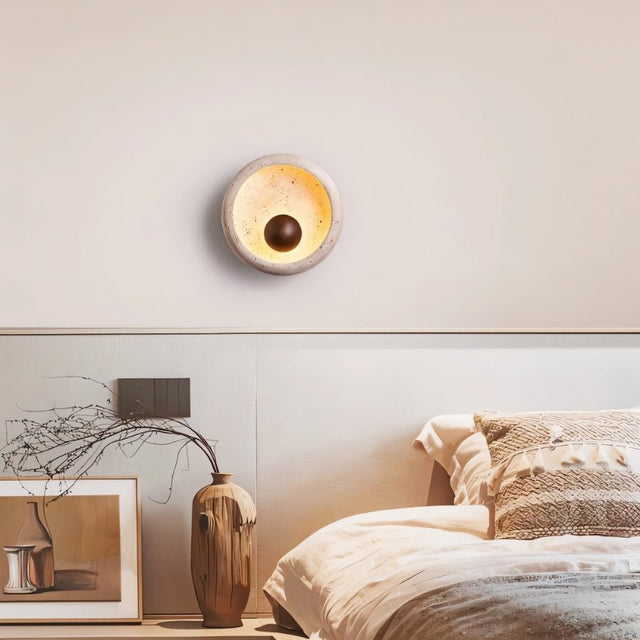 "A modern rustic wall lamp with a circular travertine frame and wooden centerpiece illuminates a contemporary bedroom."