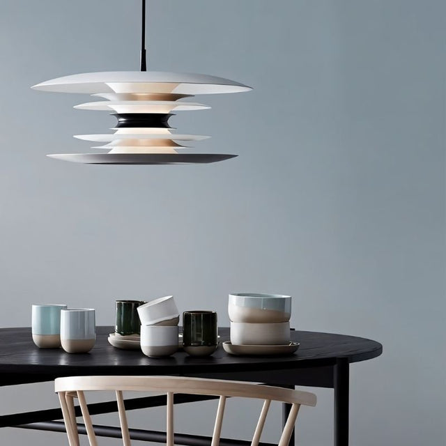 "A modern pendant light with layered discs in white and black, hanging above a dining table with cups and a chair."