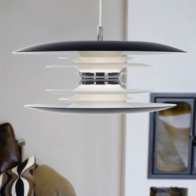 "A modern pendant light with layered discs in black and white, hanging in a living room with a patterned pillow and a fireplace."