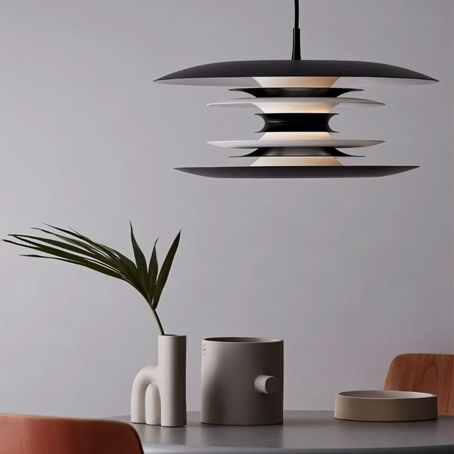 "A modern pendant light with layered discs in black and white, hanging above a table with modern decor and a plant."