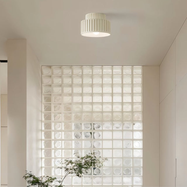 "A white ceiling light with a ribbed, cylindrical shade, shown installed on a white ceiling in a hallway with a glass block wall and a plant."