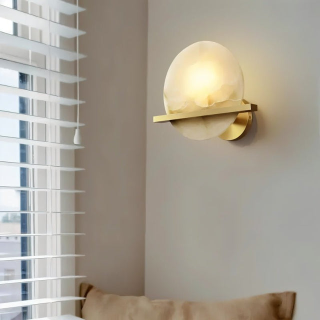 "Round alabaster wall lamp with brass frame, casting a warm light by a bedroom window."