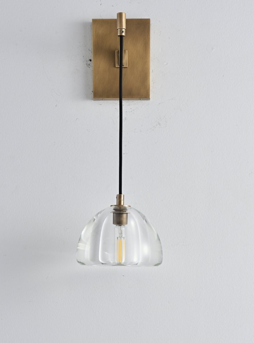 "A modern brass wall lamp with a dome-shaped clear glass shade, blending elegance and minimalist design with a curved arm and rectangular base."
