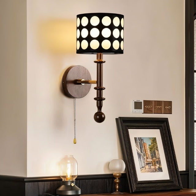"A walnut and gold wall lamp with a patterned black shade and pull chain, installed on a wall with a picture frame and other decor."