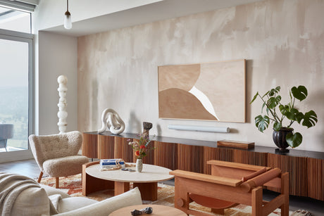 Modern living room with warm neutral tones, featuring a curved sofa, woven lounge chair, wood furniture, abstract wall art, indoor plants, and large windows overlooking a city view by the scale collective.