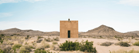 Desert landscape featuring a minimalist monolithic structure with a single dark doorway, a person seated on its roof, and rugged mountains stretching across the horizon beneath a clear blue sky.