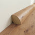 Medium-warm wood quarter round trim with a smooth, rounded edge, attached to a light wall above textured wood flooring.