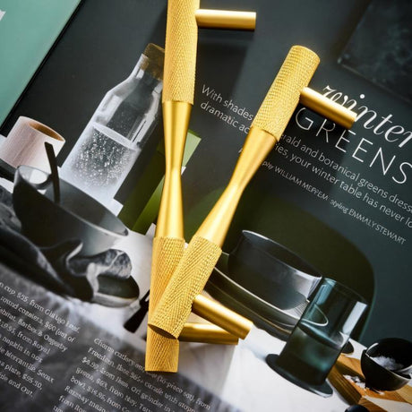 "Two brass pull bar handles with knurled detailing, styled in a lifestyle setting with a magazine and decorative objects."