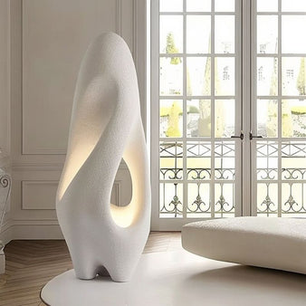 "Izbu Floor Lamp positioned by a window, highlighting its sculptural form, smooth curves, and hollow details with a soft ambient light, crafted from polystyrene."